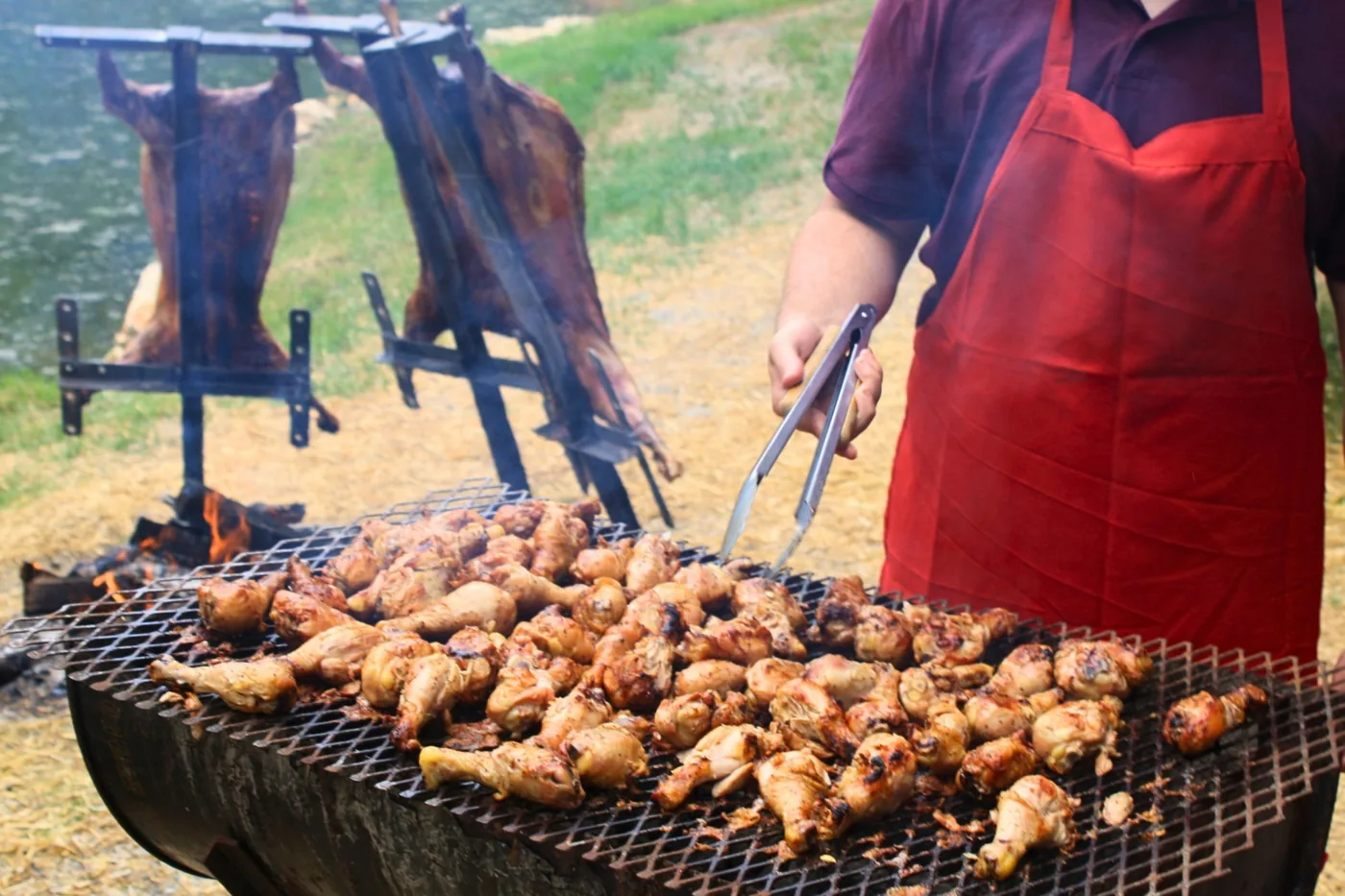 Global British Grill chef grilling chicken over open flame with whole asado roasting behind