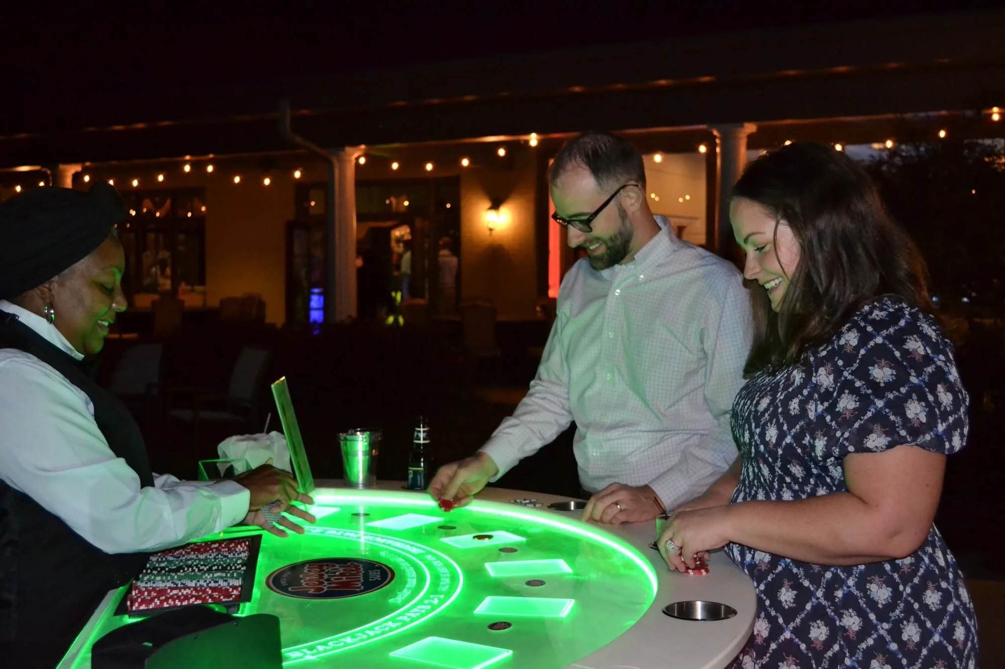 Dealer and guests at a green-lit sponsored glow table at an outdoor charity casino night